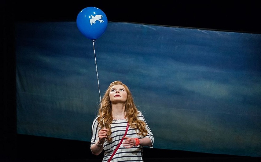 Powiększ obraz: Judyta to młoda dziewczyna, ma długie, rude włosy. Trzyma w ręku biały sznurek. Jest on przymocowany do niebieskiego balona z białym gołębiem. Judyta patrzy w górę na balon.