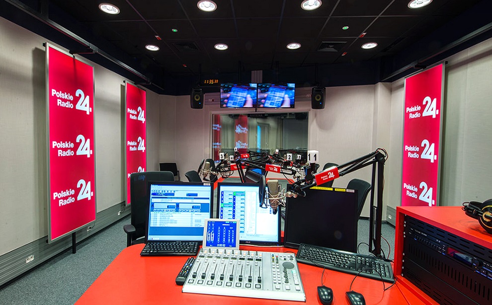 Powiększ obraz: Studio radiowe. W centrum zdjęcia biurko na którym leży konsola, za nią trzy monitory, nad nią kilka mikrofonów. Obok konsoli klawiatura i dwie myszki. Na szarych ścianach trzy czerwone podłużne bannery. Na nich białe napisy Polskie Radio 24.