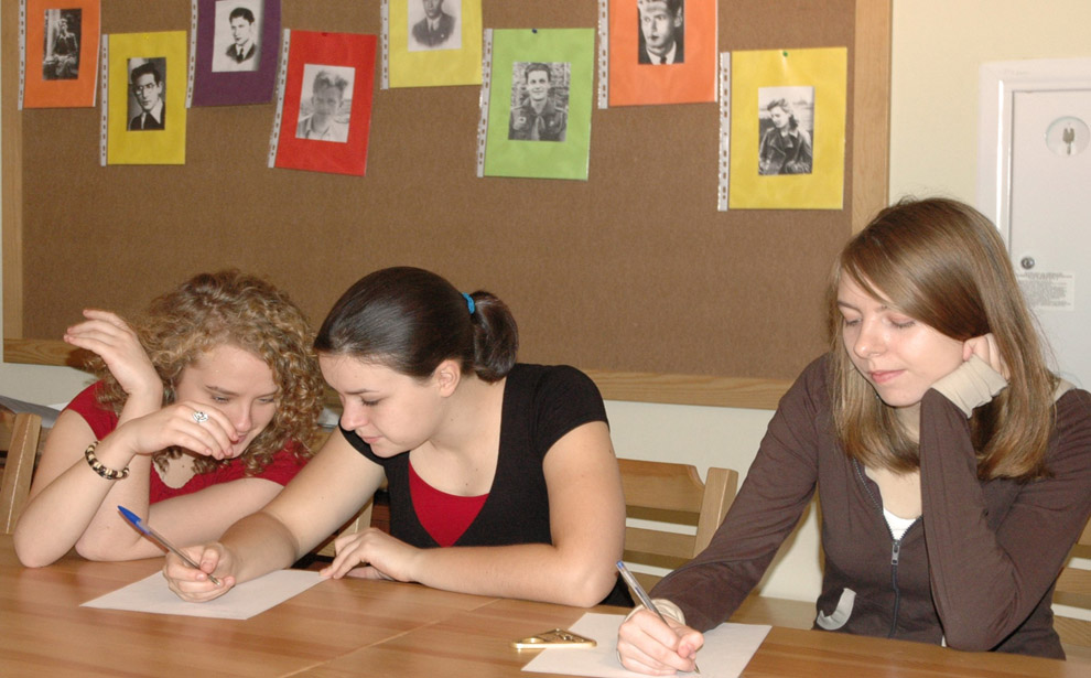 Powiększ obraz: Trzy dziewczyny siedzą przy stole, rozwiązując zadania na kartkach.  Za nimi do tablicy przypięte zdjęcia Powstańców.