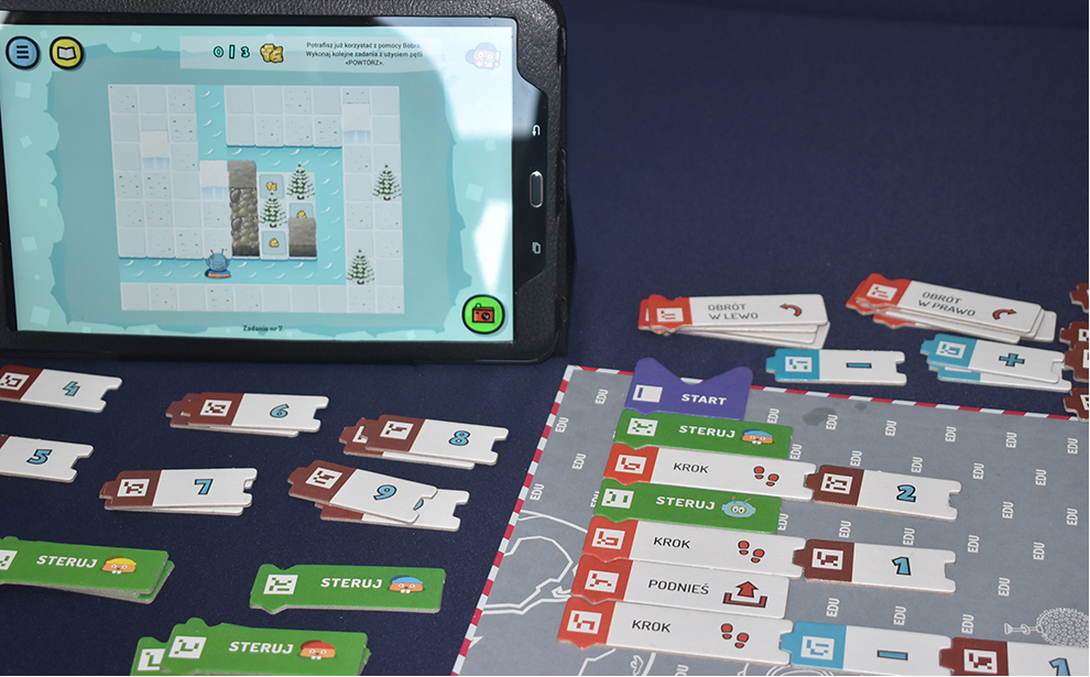 Powiększ obraz: gra edukacyjna ScottieGo! Przed ekranem komputera leżą karty do gry i papierowa plansza. Na monitorze elektroniczna wersja planszy.