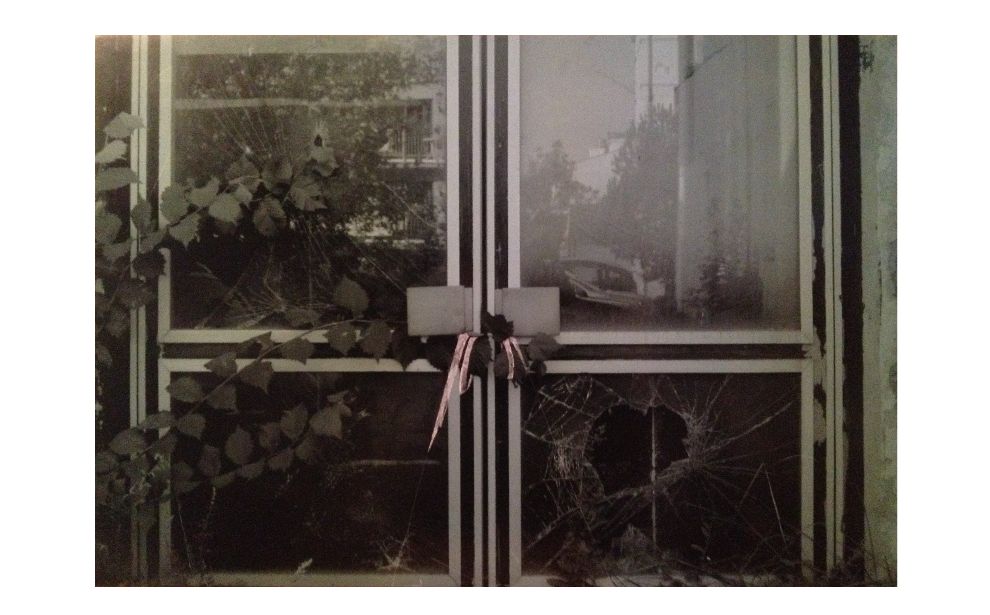 Powiększ obraz: Praca Bianki Rolando z cyklu “Wschody”. Czarno-biała fotografia rozbitych, szklanych drzwi. W centrum, pod klamką fragmenty obrazu zostały wydrapane przez artystkę.