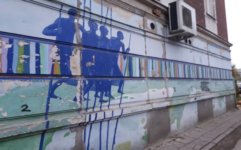 Powiększ obraz: Praski Street Art. Na ścianie kamienicy niebieski mural. Przedstawia cztery postacie z dzidami.